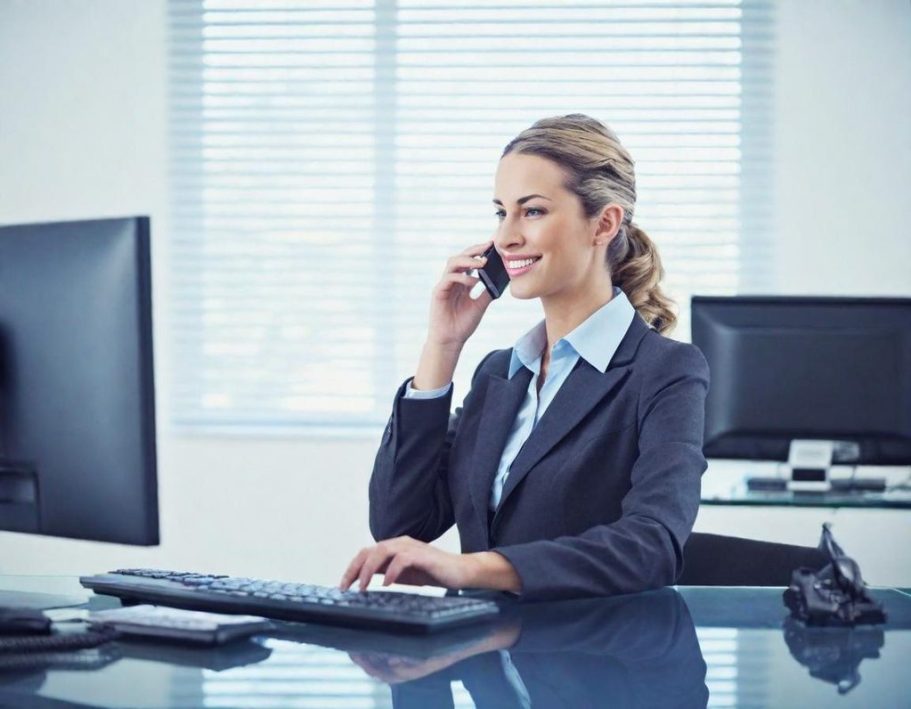 Femme en costume, souriante, au téléphone, assise devant un ordinateur.